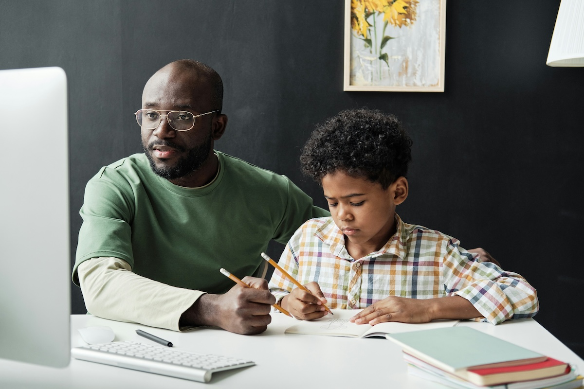 A man looks on at a computer screen with pencil in hand helping a child with homework