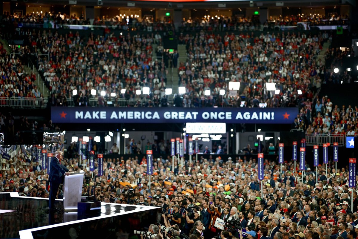 A large stadium crowd is assembled and listening to Donald Trump speak on stage. A big sign reads Make America Great Once Again!
