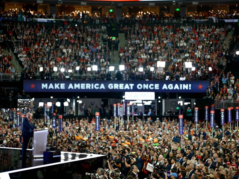 A large stadium crowd is assembled and listening to Donald Trump speak on stage. A big sign reads Make America Great Once Again!