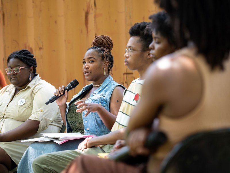 A photo of a Black woman with her hair in a bun speaking into a microphone while sitting on a panel.