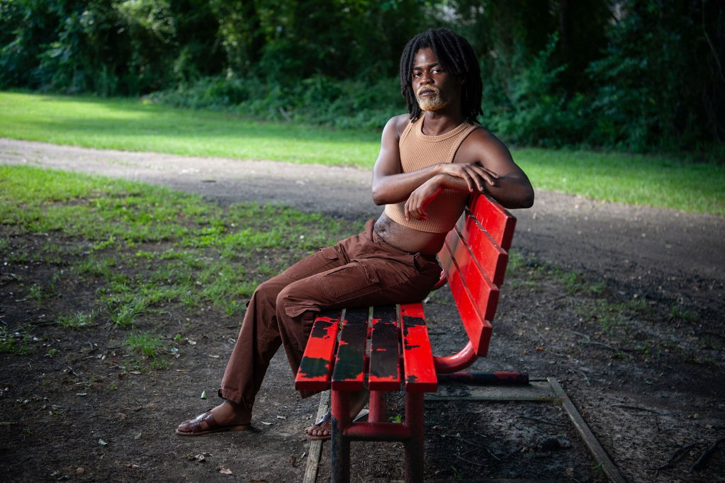 A photo of a Black person with black hair and a blond beard sitting on a red park bench while looking at the camera.