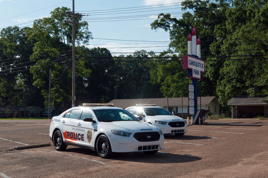 A photo of two white police cars in a parking lot in the middle of the day. Behind the cars is a strip mall sign reading “Candlestick Park” with three candlestick figures.
