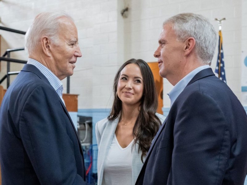Two men in suits look at each other and shake hands as a woman smiles on