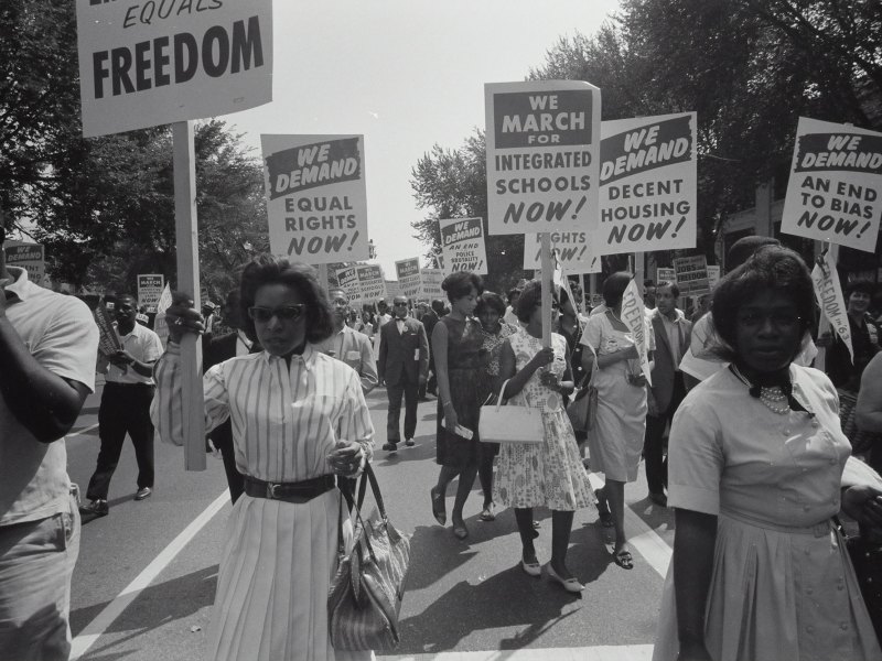 A group of black people march down the road with signs that ready We Demand Equal Rights Now!