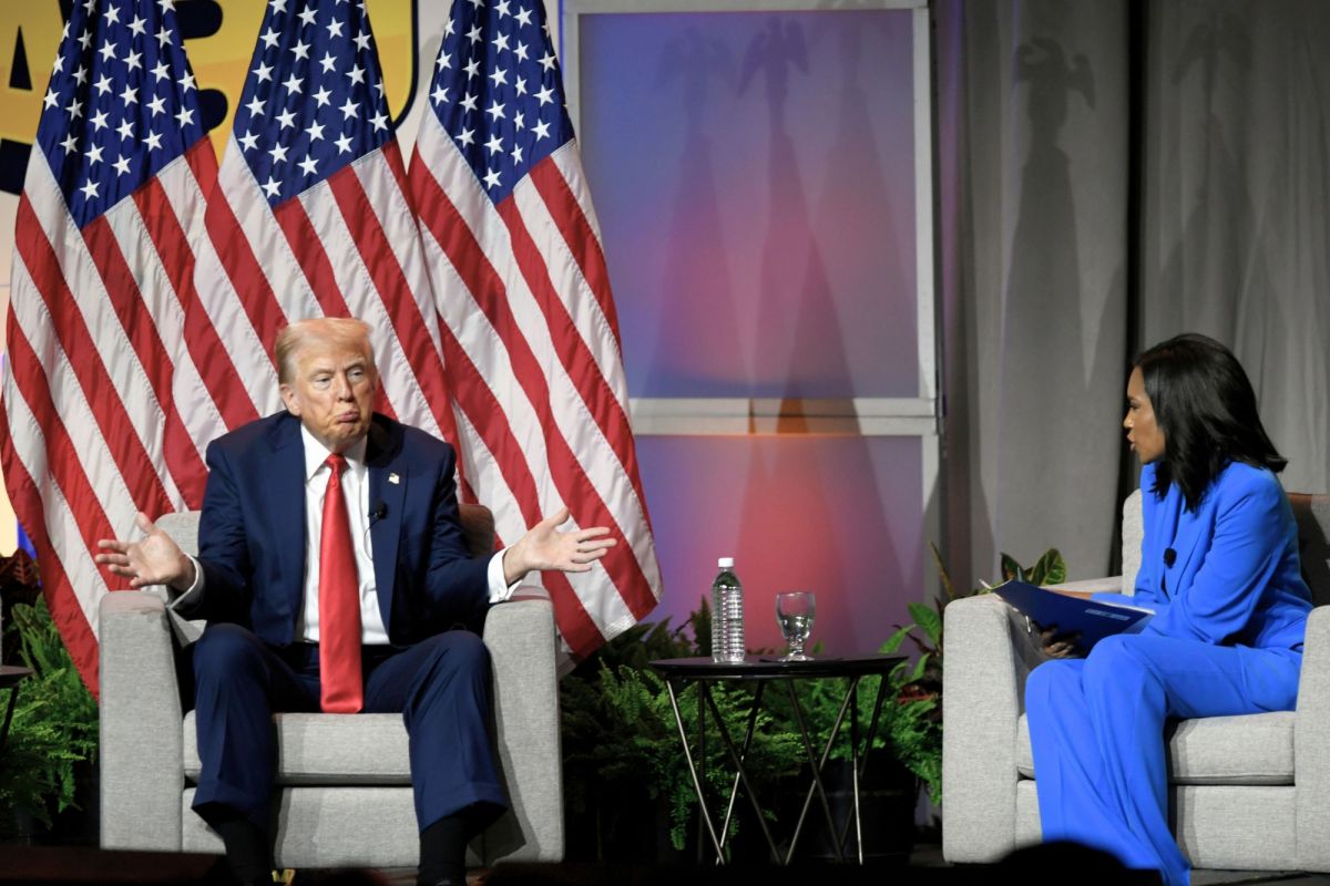 A man in a suit sits in a chair backed by three US Flags, shrugging while looking at a woman sitting in the opposite seat.