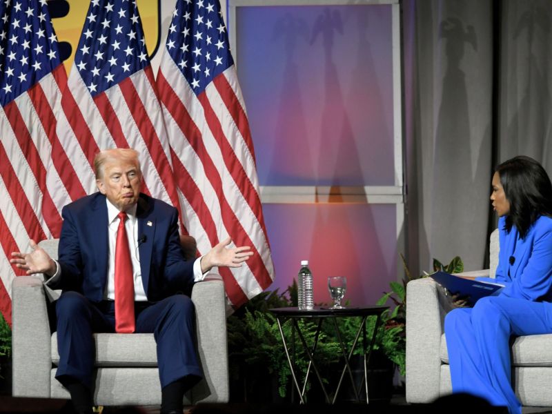 A man in a suit sits in a chair backed by three US Flags, shrugging while looking at a woman sitting in the opposite seat.