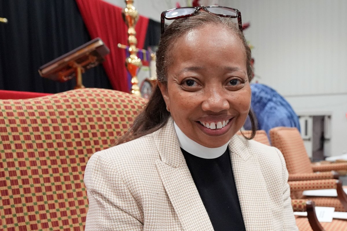 A woman with a bishop collar sits in a large checkered chair, a cross visible on the stage behind her