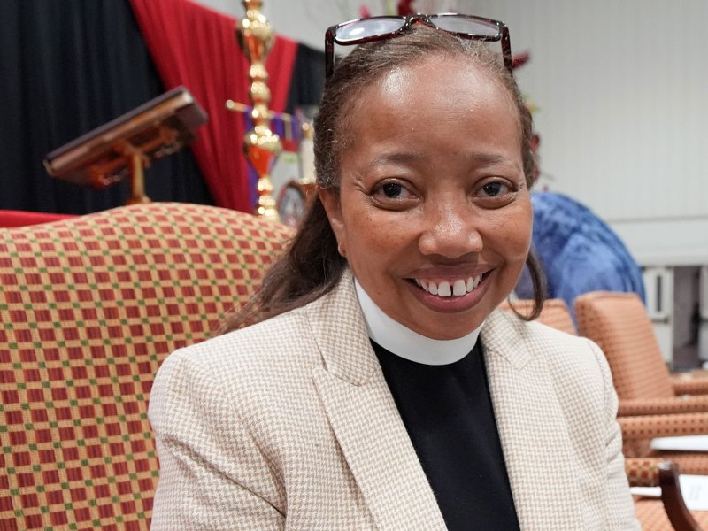 A woman with a bishop collar sits in a large checkered chair, a cross visible on the stage behind her