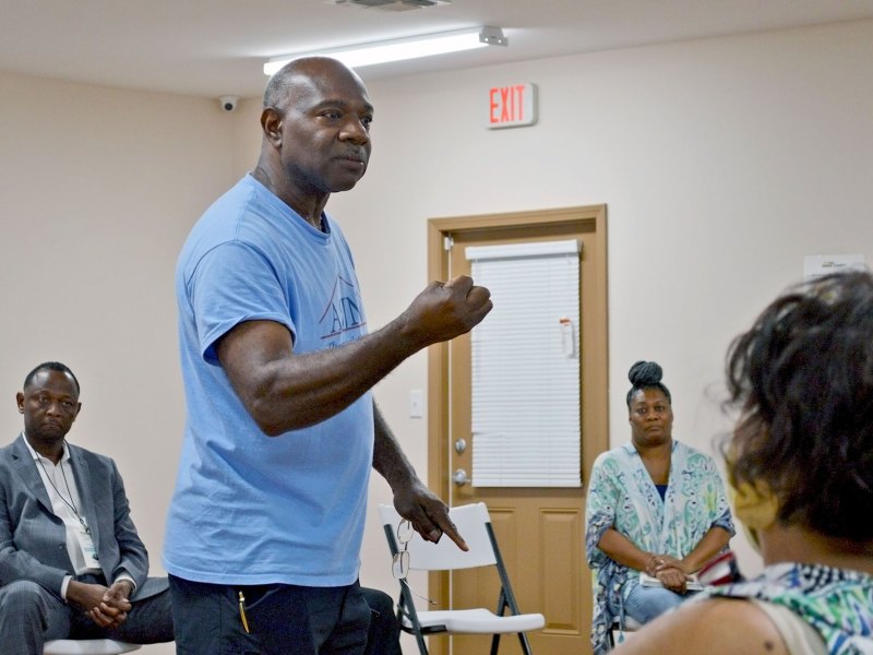 A man in a blue tshirt speaks to a seated audience inside a room.