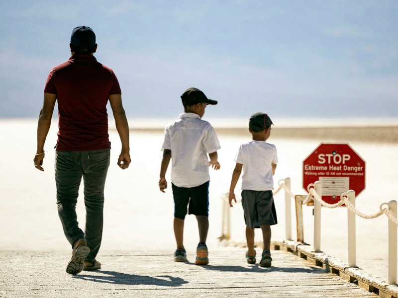 An adult and two children go along a boardwalk in the sand. A red stop sign reads STOP Extreme Heat Danger