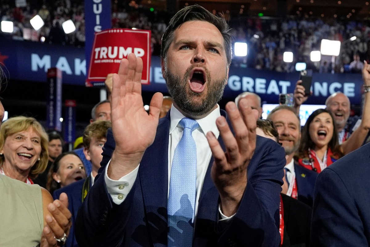 A man in a suit claps while yelling something. A crowd with signs for Trump stand behind him