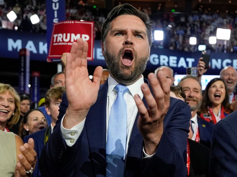 A man in a suit claps while yelling something. A crowd with signs for Trump stand behind him