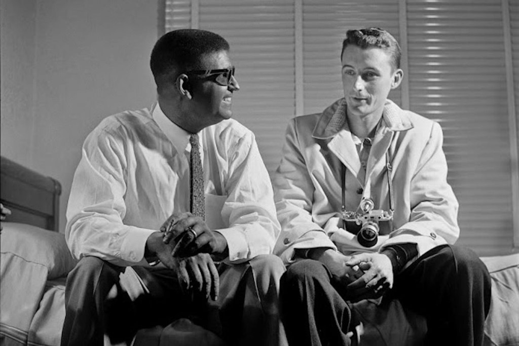 Smiling Black man wearing a tie sits on a bed while talking to a young white man with a camera hanging from his neck