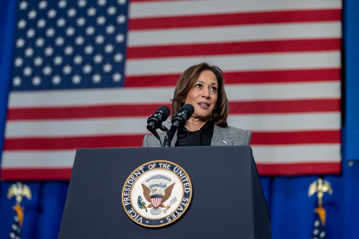 A woman stands behind a podium labeled Vice President of the United States. A large US Flag covers the wall behind her.
