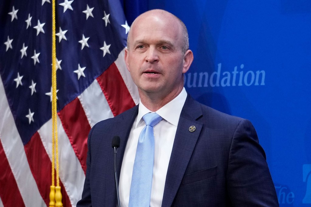 A man in a suit stands before a US Flag and a blue background that has a cut off "Foundation" written on it