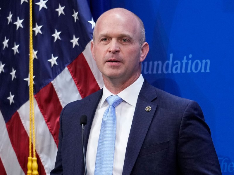 A man in a suit stands before a US Flag and a blue background that has a cut off "Foundation" written on it