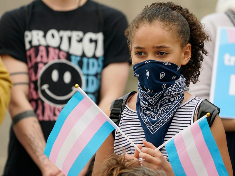 A young girl with her face partially covered holds two trans flags