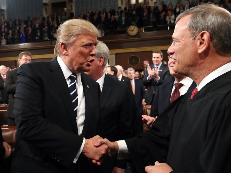 Two men shake hands as many people in the room behind them stand and clap
