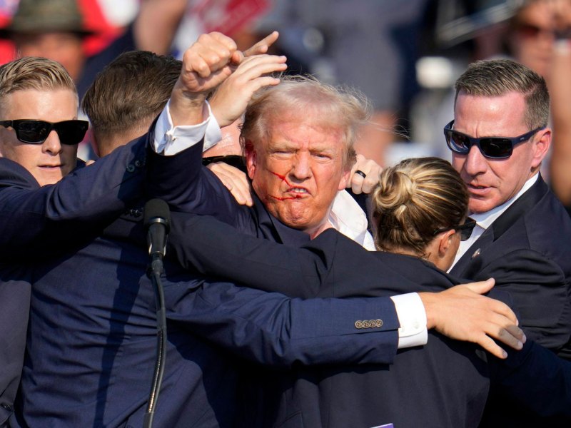 Four people protectively put their bodies around one older man who has his arm raised in a fist