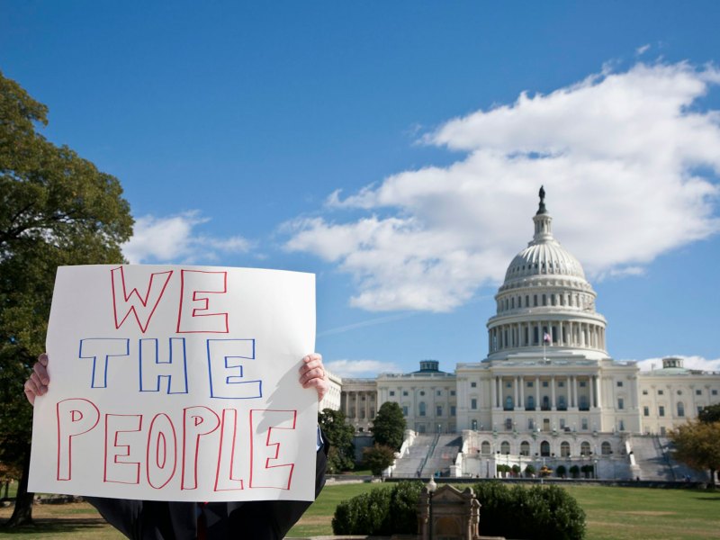 A person holds a handwritten sign that says "We the People" while they stand in front a capitol building