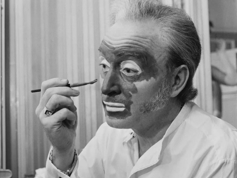 A black and white photo of a man applying dark makeup to his face