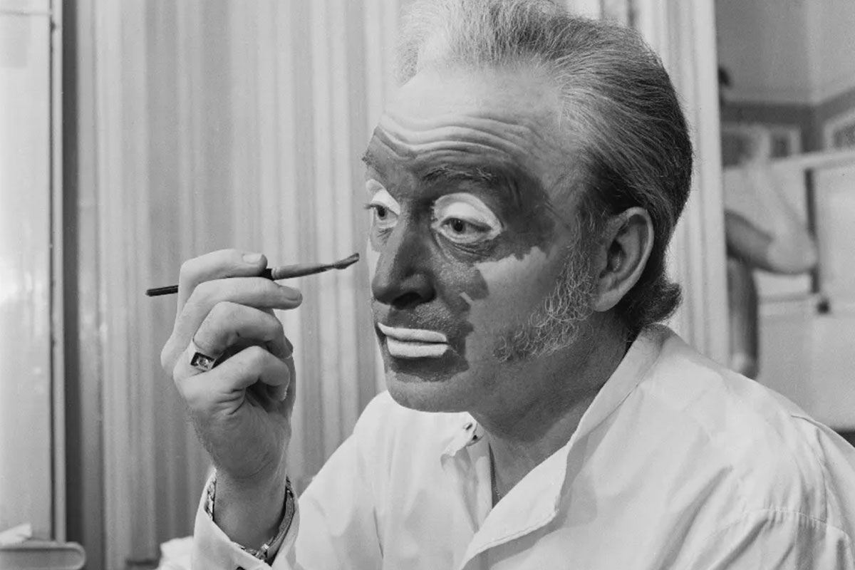 A black and white photo of a man applying dark makeup to his face