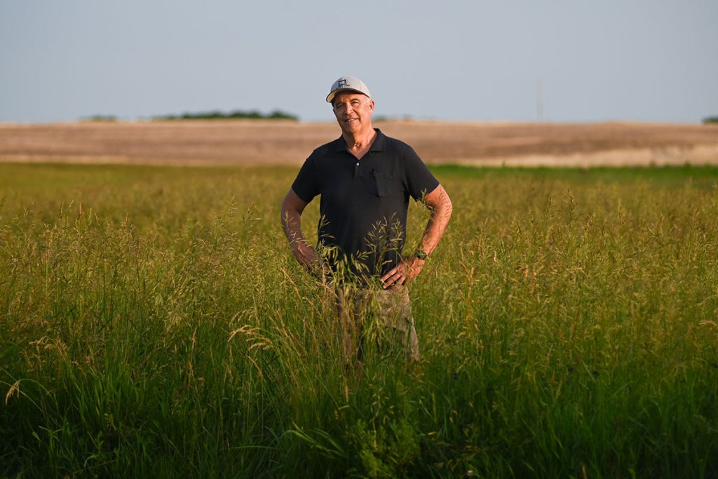 A man in a black shirt and white ballcap stands in a field that's grown waist-high