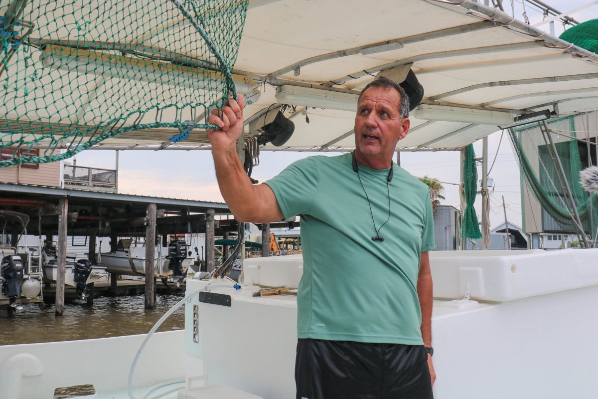 A man in a green short holds onto a hanging fishing net on his boat