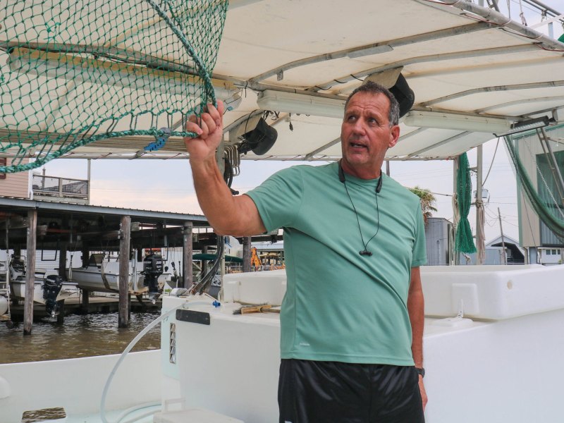 A man in a green short holds onto a hanging fishing net on his boat