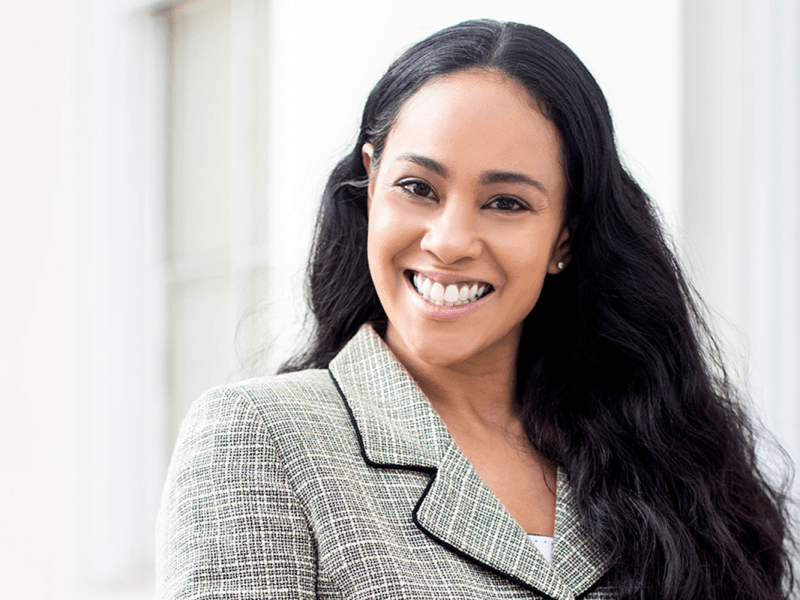 A photo of a smiling woman with long black hair and grey suit top standing outside