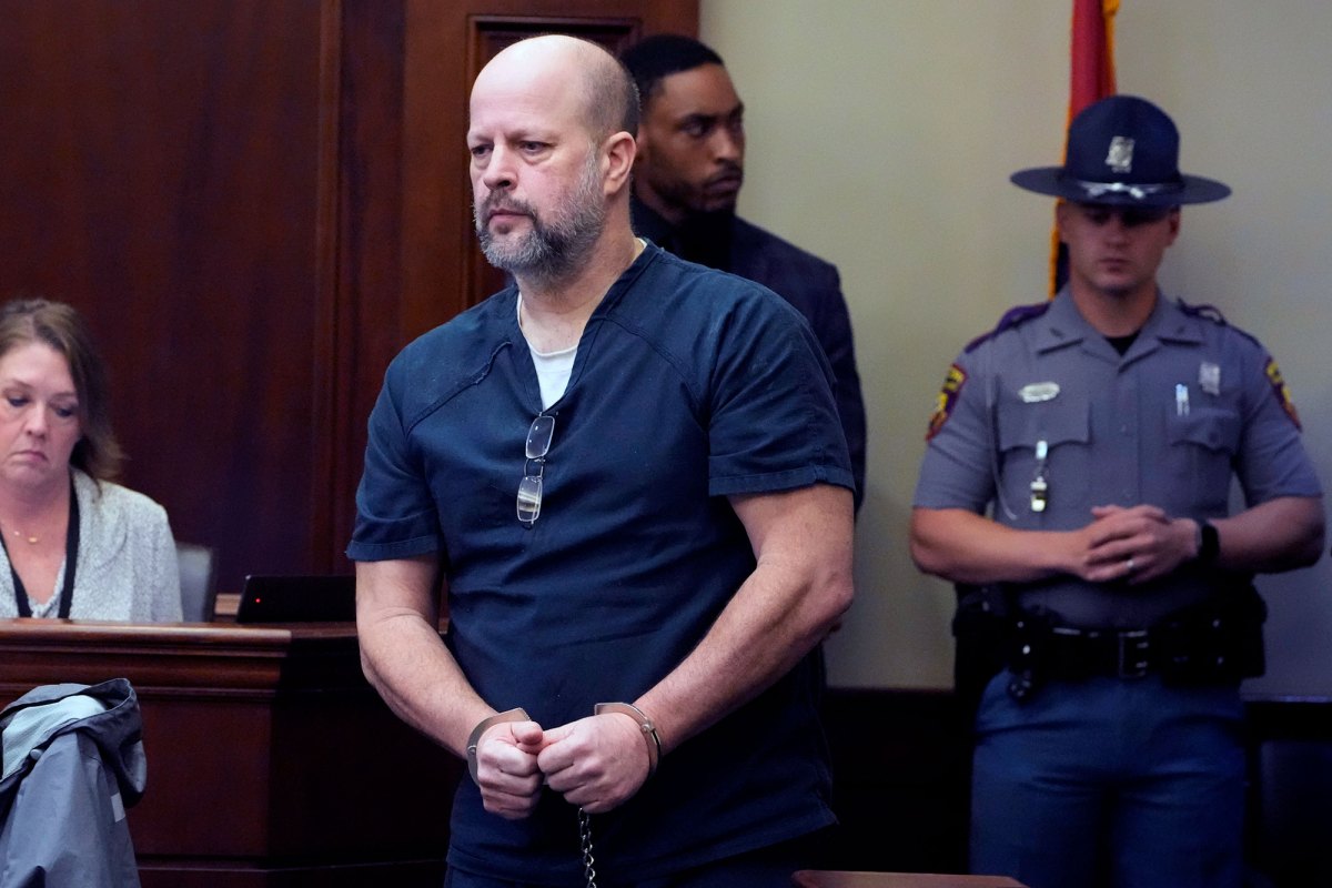 A bald man with a beard walks through a courtroom while in handcuffs