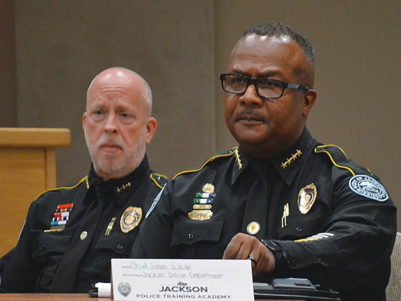 Two police officers sit at a table at a discussion. A sign before them reads Jackson Police Training Academy