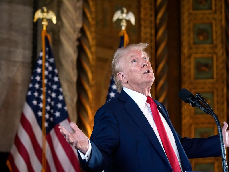 Donald Trump, wearing a dark blue suit and red tie, speaks at a mic with arms held wide and face looking up at the ceiling. Behind him are two US flags inside a golden opulent room.