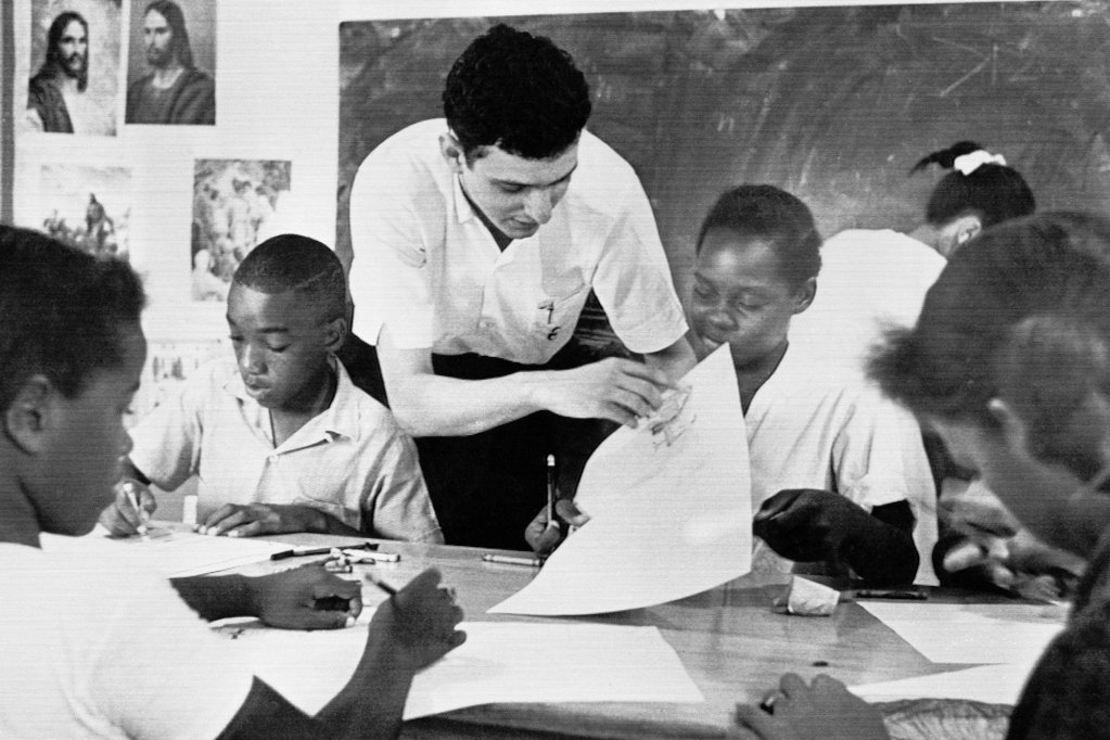 A black and white photo showing a white teacher leaning over and helping a table full of black students in the classroom