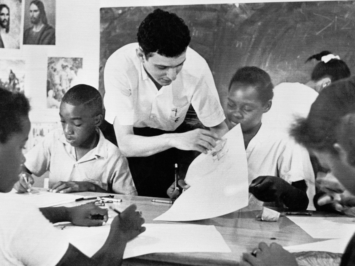 A black and white photo showing a white teacher leaning over and helping a table full of black students in the classroom