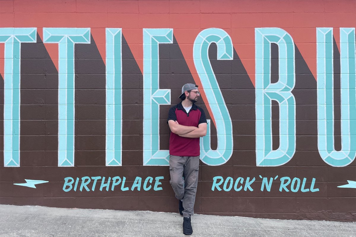 A man wearing a black and red polo shirt paired with a backwards grey baseball cap and grey jeans leans casually against a wall painted as a mural. The words are cut off but it reads Hattiesburg, Birthplace of Rock 'n Roll