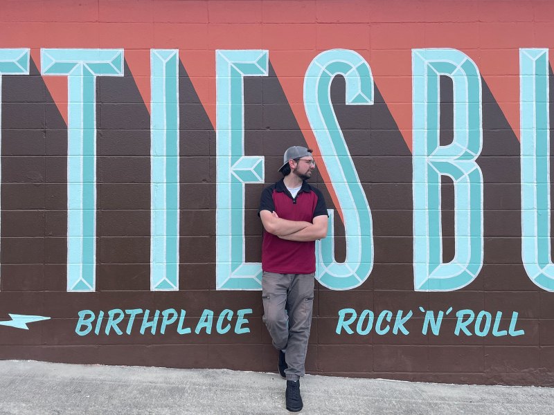 A man wearing a black and red polo shirt paired with a backwards grey baseball cap and grey jeans leans casually against a wall painted as a mural. The words are cut off but it reads Hattiesburg, Birthplace of Rock 'n Roll