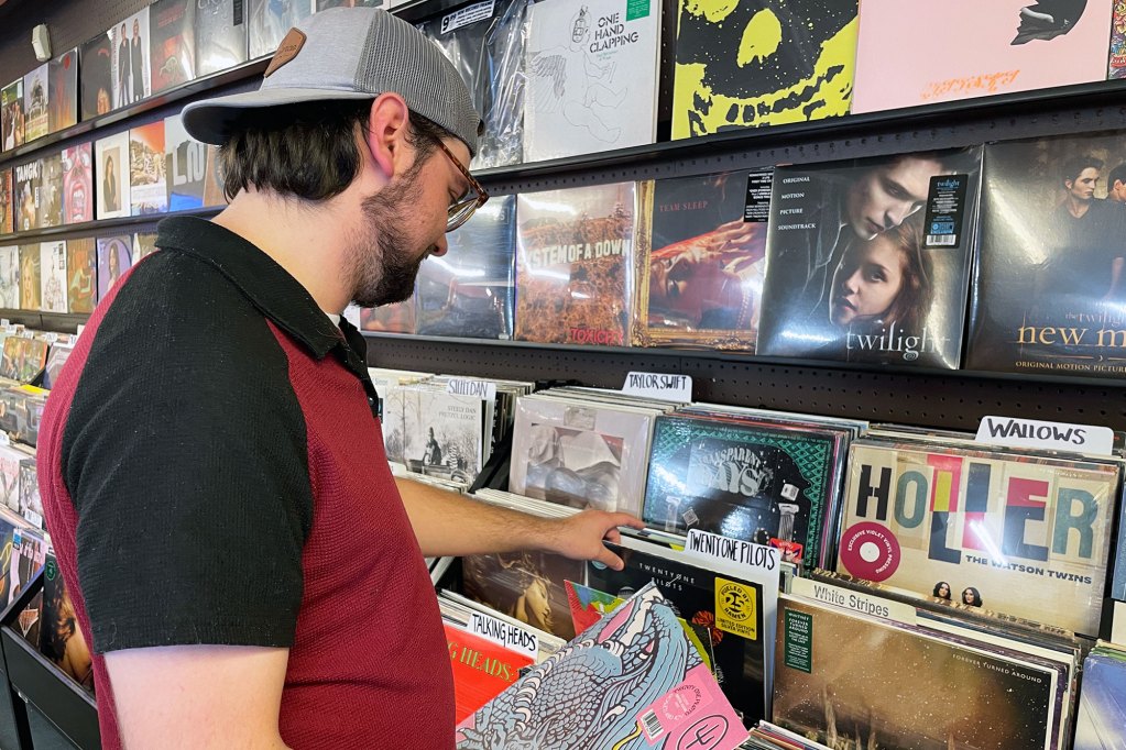 A man wearing a black and red polo shirt paired with a backwards grey baseball cap peruses a colorful wall of records