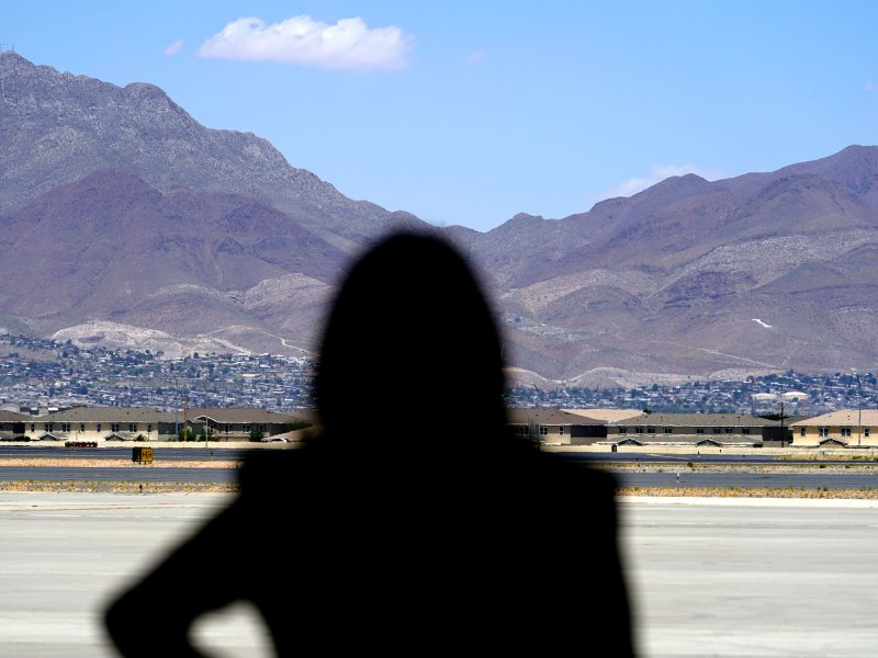 A silhoutte of a woman looking onto small buildings with a town further in the background, and tall mountains behind everything