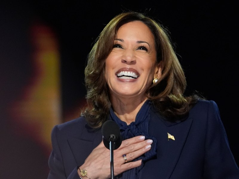 A woman in a dark blue suit smiles at the mic with one hand on her chest