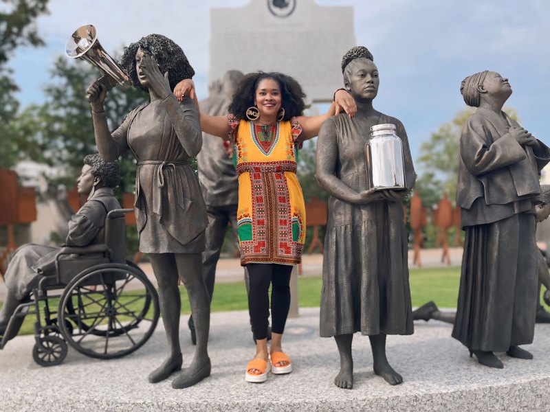 A photo of a woman in orange toned clothes standing between four metal statues of women