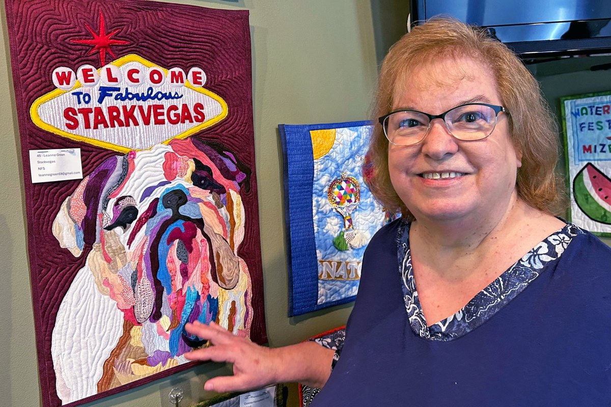 A woman stands in front of a quilted art piece depicting a bulldog and a sign that reads "Welcome to Fabulous Starkvegas"