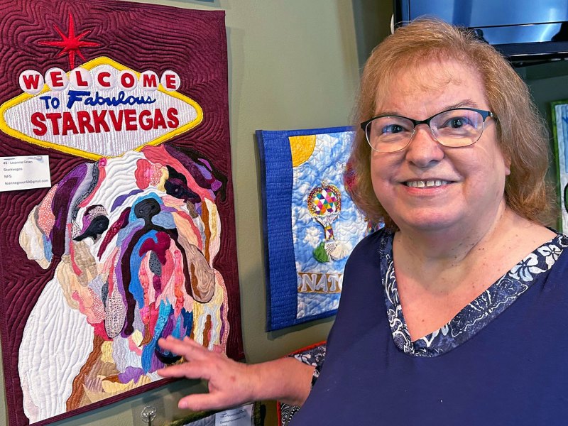 A woman stands in front of a quilted art piece depicting a bulldog and a sign that reads "Welcome to Fabulous Starkvegas"