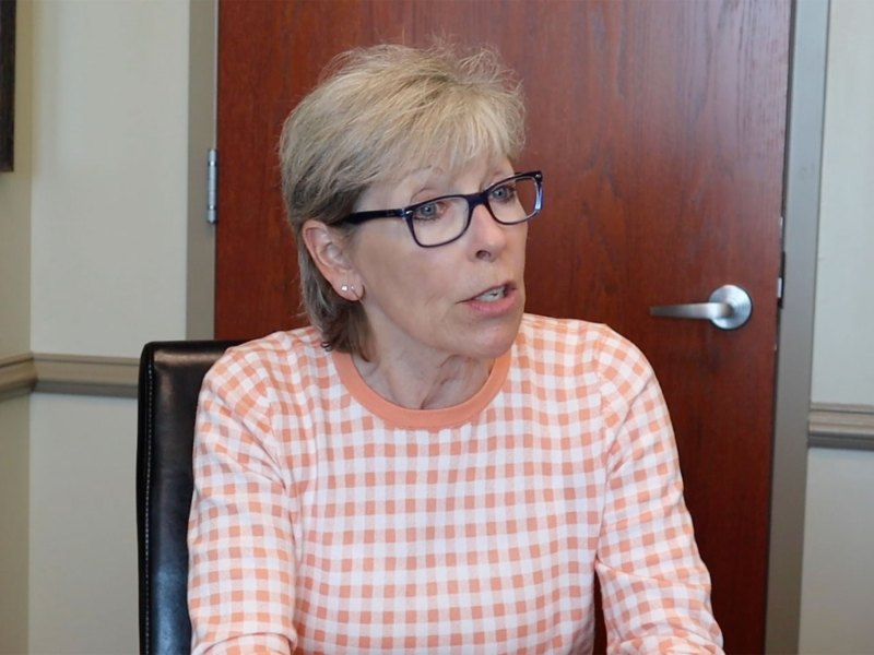 A woman with short blonde hair and black glasses, wearing a white and orange checkered sweater sits in and speaks inside an office