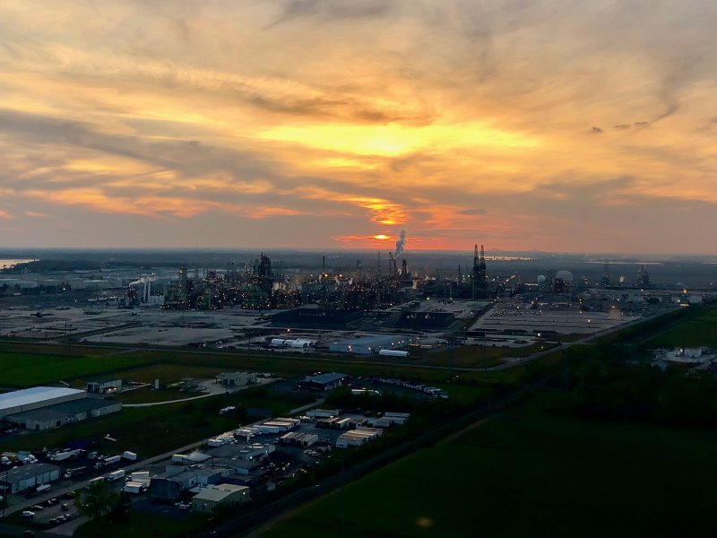 An aerial view of a refinery at sunrise, large stacks can be seen in the distance emitting plumes of smoke