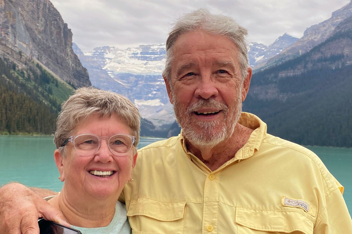 A woman and a taller man pose for a photo with a lake and snow capped mountains behind them