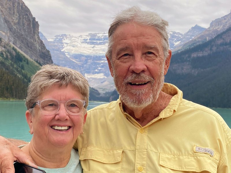 A woman and a taller man pose for a photo with a lake and snow capped mountains behind them