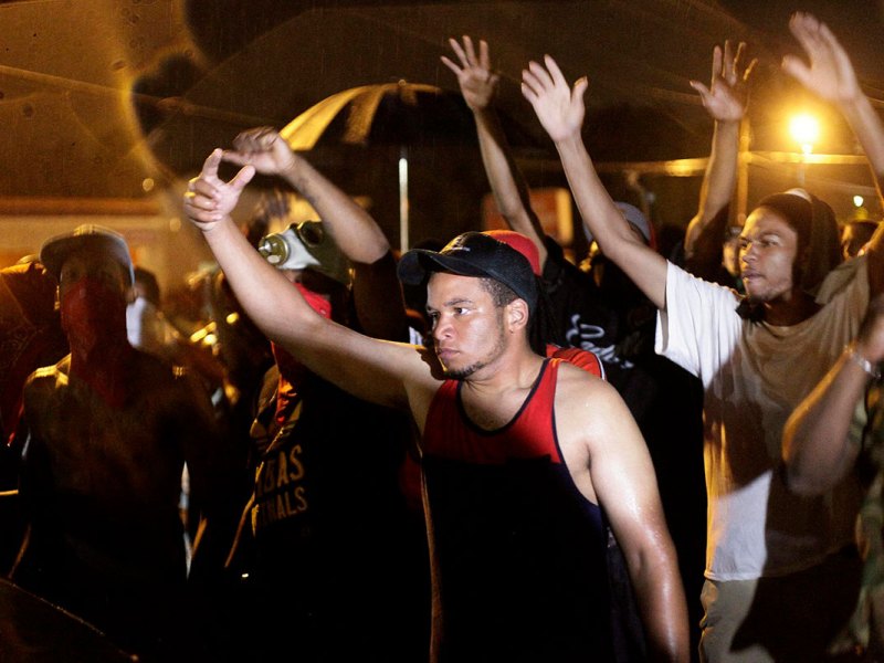A crowd stands with arms raised in the night, lit with yellow lights
