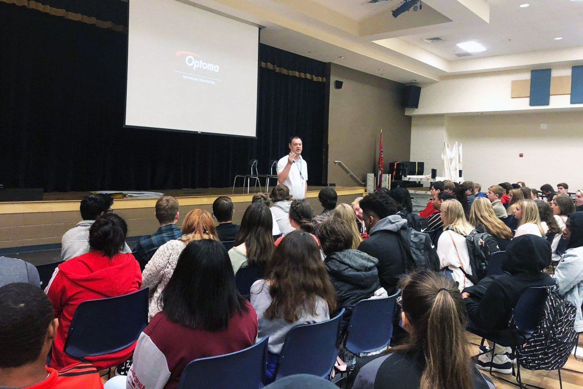 A man gives a presentation in front of rows of students in a school auditorium