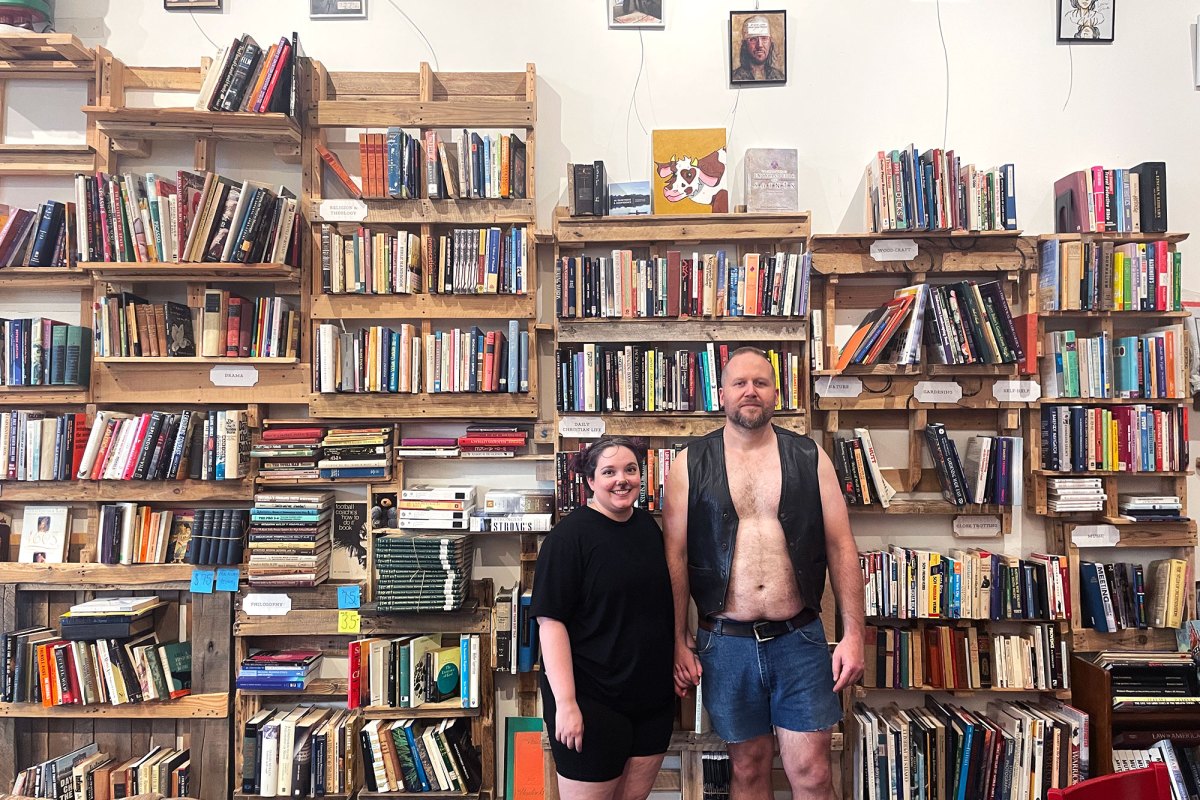 Two people stand in front of a very busy wall of bookshelves. The man on the right is taller, wearing an open black vest with no shirt and denim shorts. The woman on the left is shorter, wearing all black and light cat makeup